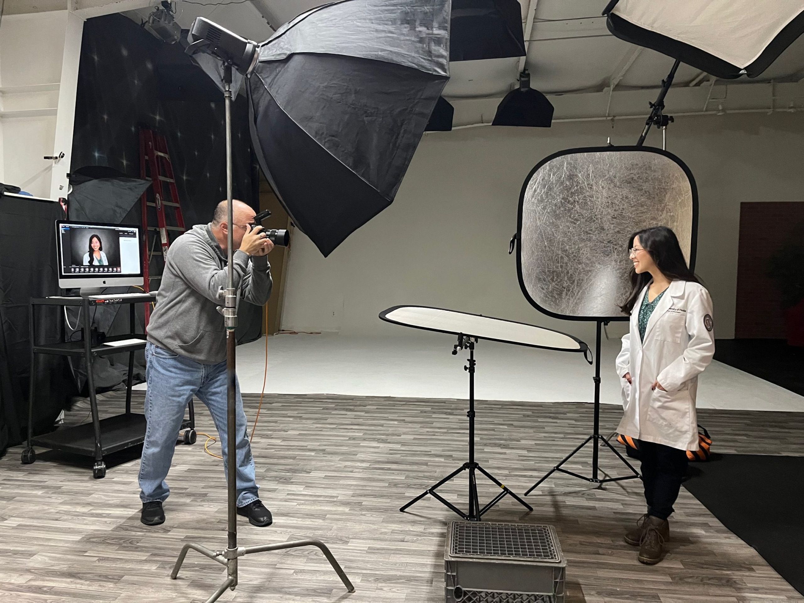 A woman wearing a white lab coat, stands in a photography studio. A photographer, who appears to be a man with a shaved head and a gray sweatshirt, is taking their picture. The studio features various lighting equipment, including a large black softbox on a stand, a silver reflector, and a smaller white reflector. A computer monitor on a stand is visible in the background, displaying a portrait. The floor is a light-colored wood laminate.