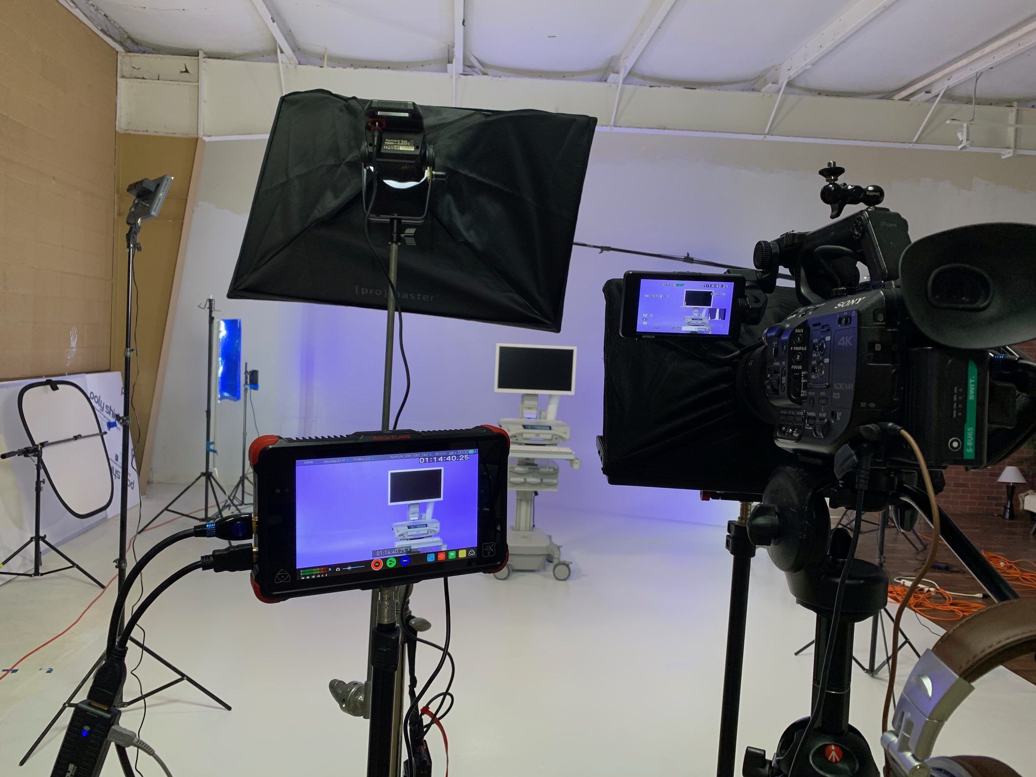 Behind-the-scenes view of a professional video production set in a studio with a white cyclorama. The image shows a professional camera, multiple external monitors, and a large softbox light focused on filming medical equipment.