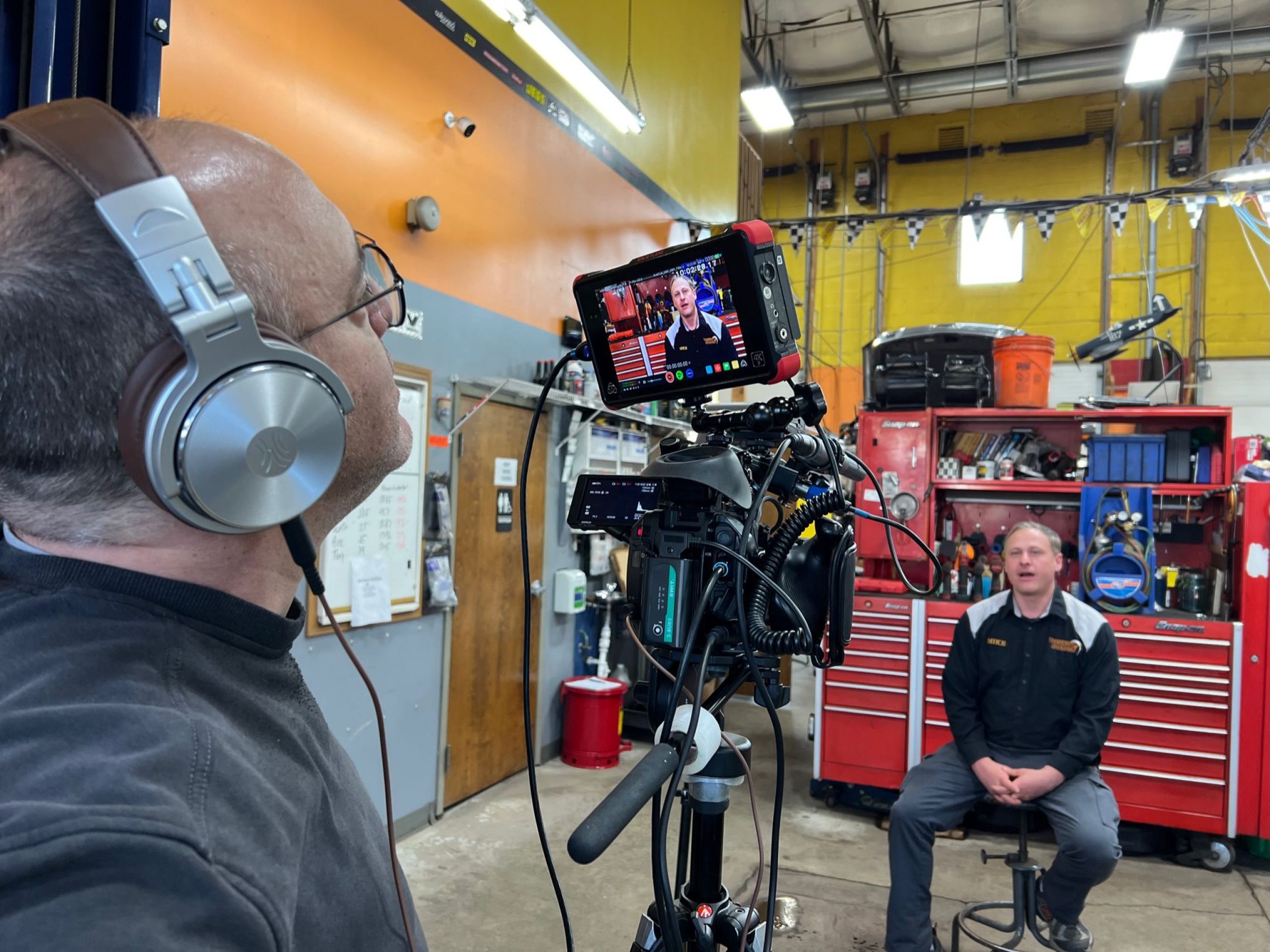A man with glasses and headphones faces away from the camera, focusing on a video monitor attached to a professional video camera. The monitor shows a man speaking. In the background, a man sits in a chair. The setting appears to be an auto repair shop with tools and equipment visible.
