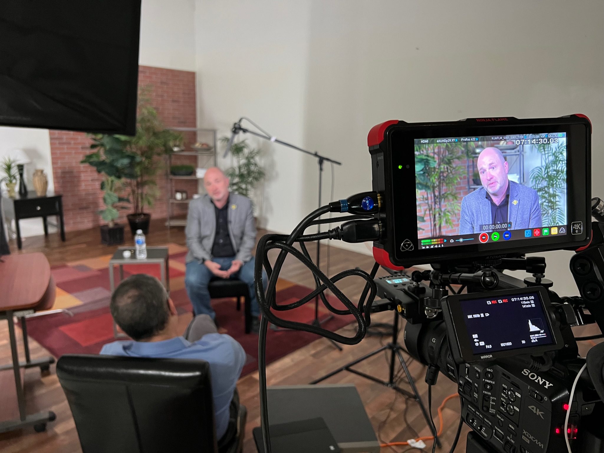 Camera recording interview with man in grey jacket on monitor, second man in blue shirt facing away, studio setting with plants and red rug.
