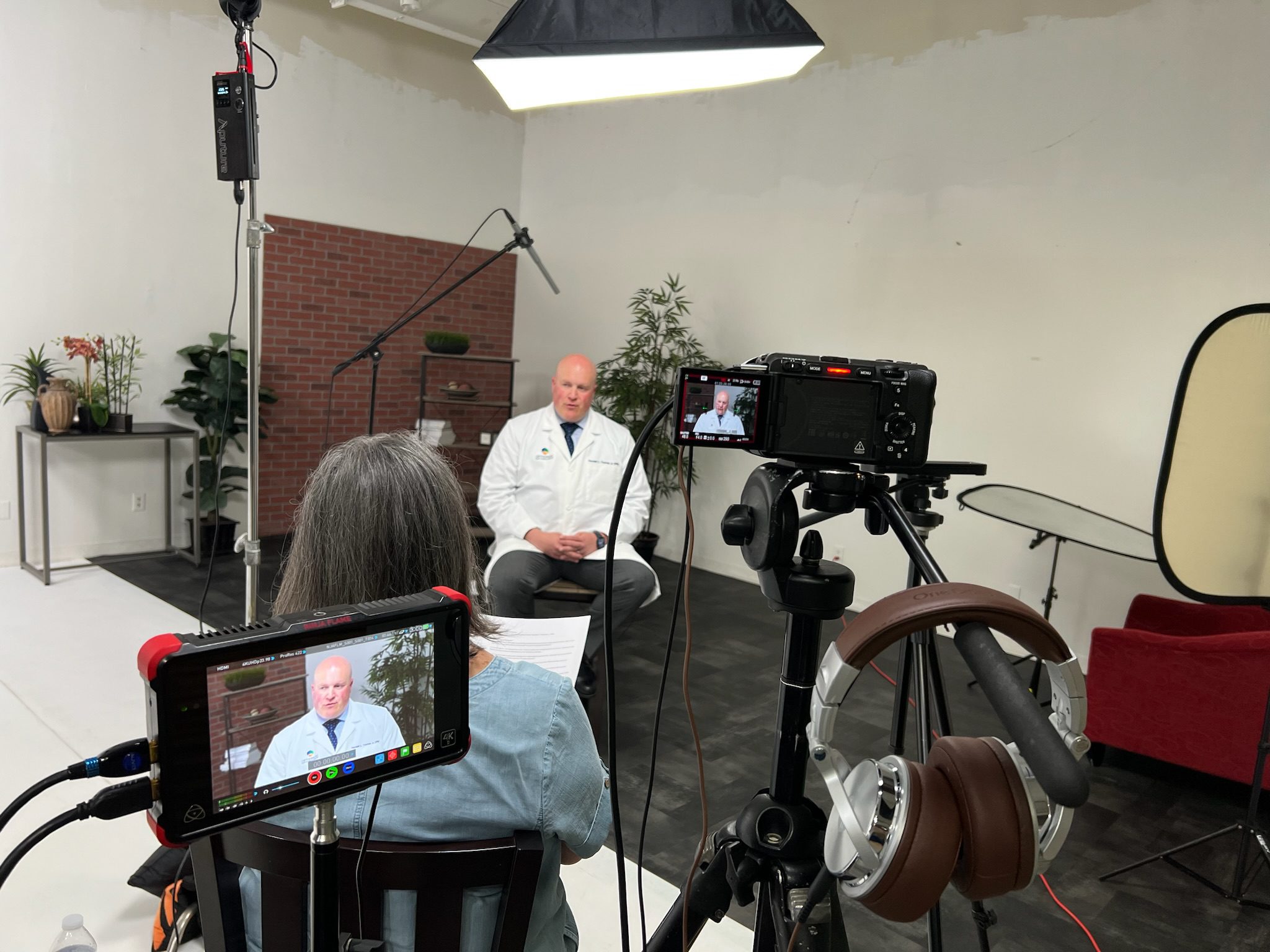 An over-the-shoulder, behind-the-scenes shot of a professional video interview with a doctor in a white lab coat. The studio setup includes a camera, external monitor, boom microphone, and professional lighting.